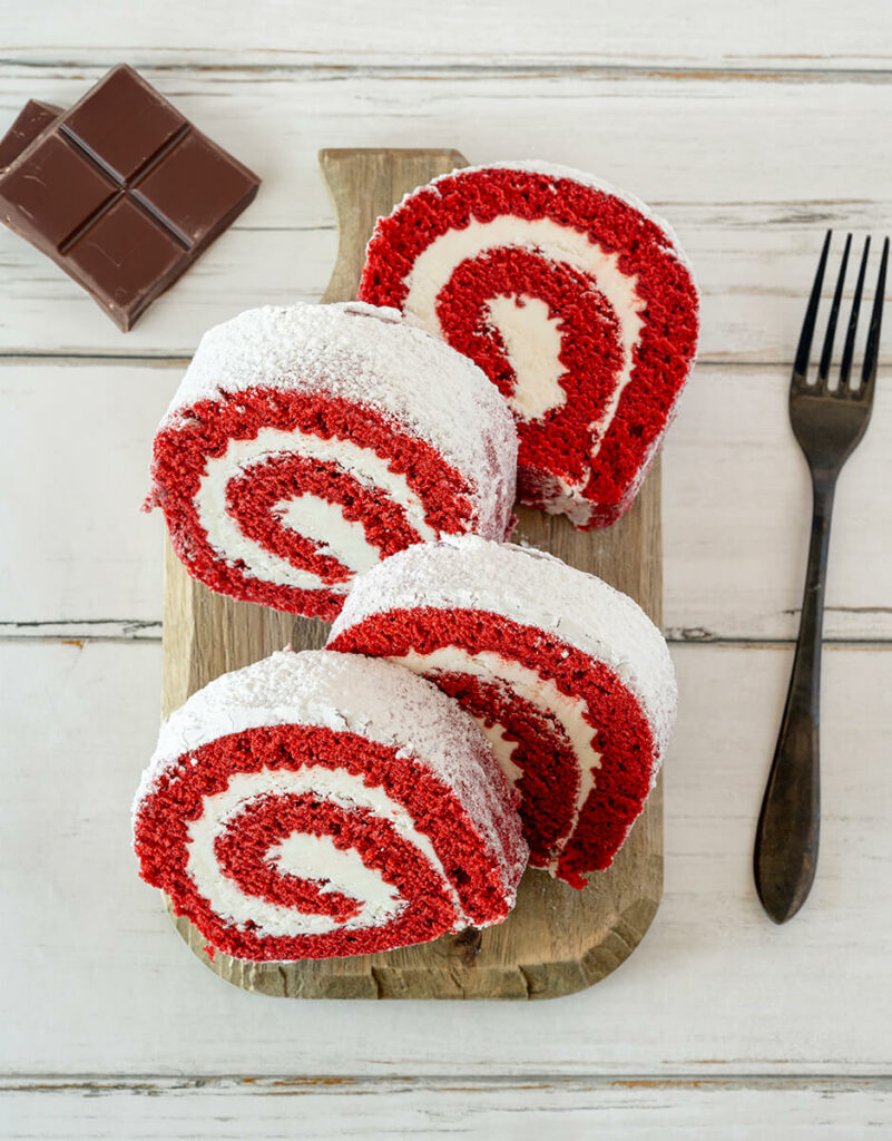 Slices of Red Velvet Cake Roll on cutting board with chocolate pieces on white wooden background