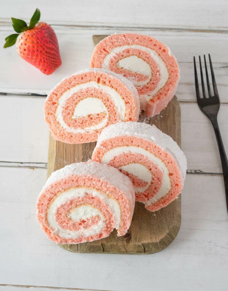 Slices of Strawberries & Cream Cake Roll on cutting board with a full strawberry on white wooden background