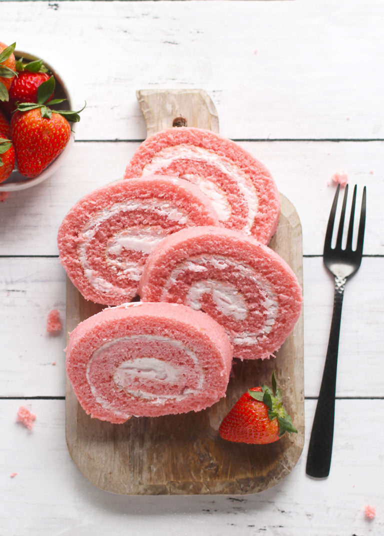 Slices of Strawberries & Cream Cake Roll with Strawberry Flavored Butter Cream frosting arranged on a wooden cutting board next to a fork and a bowl of fresh strawberries.