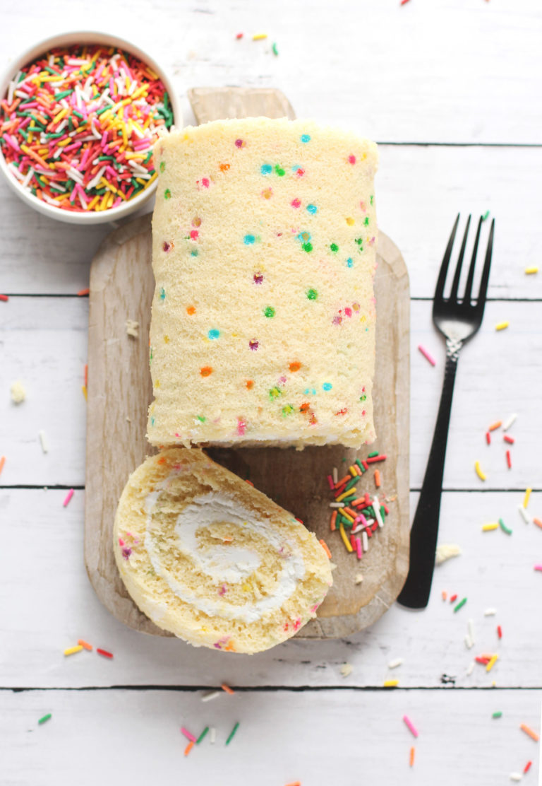 Slice of Confetti Cake Roll with Cream Cheese Frosting on wooden cutting board with a bowl of sprinkles nearby.
